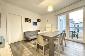 A modern living room with a grey sofa, a wooden dining table, and a large window offering a view of the outside.