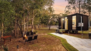 Un espace extérieur accueillant, aménagé avec des sièges confortables et un foyer, entouré d’arbres et d’un petit bâtiment dont la porte est ouverte.
