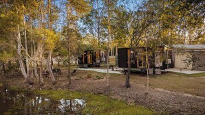 A rustic cabin with a small deck and outdoor seating area, surrounded by trees and a pond. - JJJ's Ranch Farm 1 by Tiny Away (South Kolan)