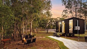 A cozy outdoor seating area with a fire pit, surrounded by trees and a small building with an open door. - JJJ's Ranch Farm 1 by Tiny Away (South Kolan)