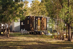 Una casa móvil con techo plano, sobre un remolque, rodeada de árboles y césped.