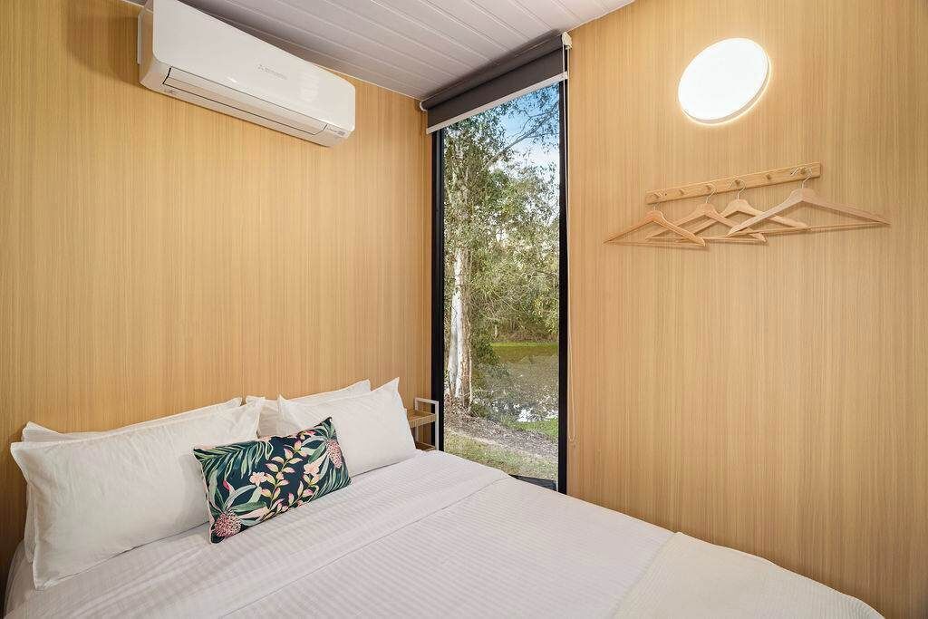Ein Schlafzimmer mit einem Bett, Kissen, einer Klimaanlage, einer Holzwand, einem Fenster und einer Wandleuchte.