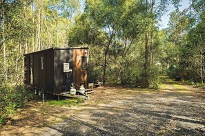 A small, dark-colored RV-style cabin with a water heater and propane tanks in a forested area.