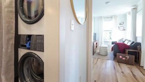 A laundry room with a washing machine and dryer, a mirror, and a hallway leading to a living area with a sofa and a table.
