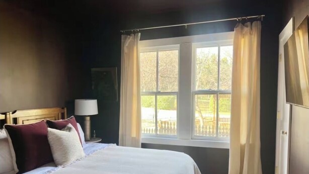 A bedroom with a large bed, a window with curtains, a nightstand with a lamp, and a view of the outside.