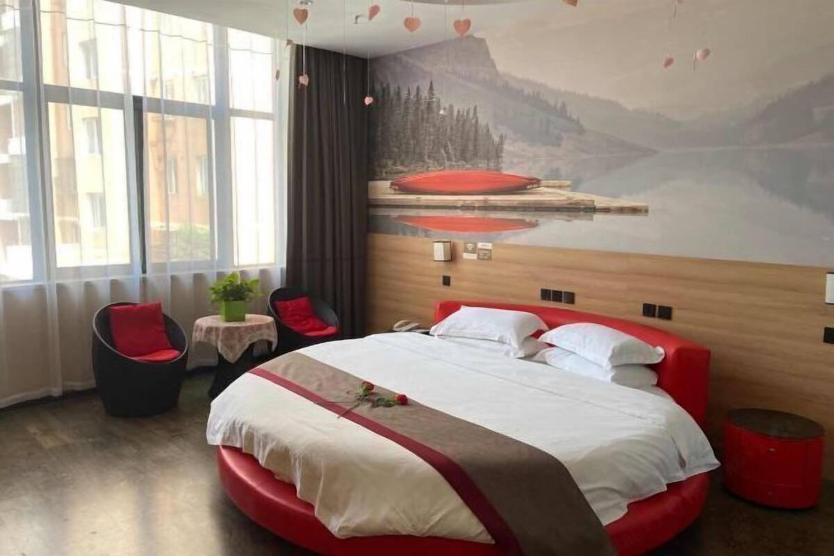 A hotel room with a large bed, a view of a mountain landscape, a small table with a plant, and a red chair.