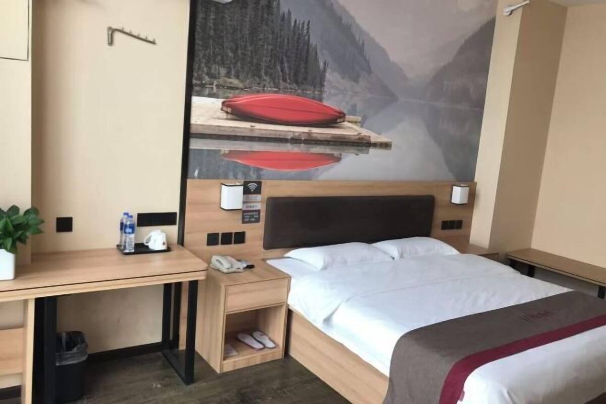 A hotel room with a bed, a desk, a chair, a small shelf, a trash bin, a potted plant, and a wall-mounted phone.