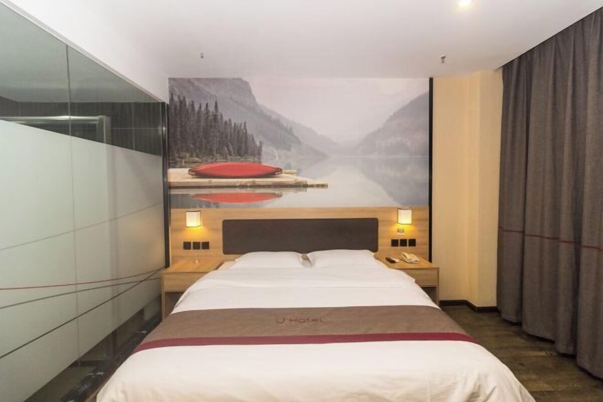 A hotel room with a bed, nightstands, a telephone, a painting of a mountain landscape, and a window with curtains.