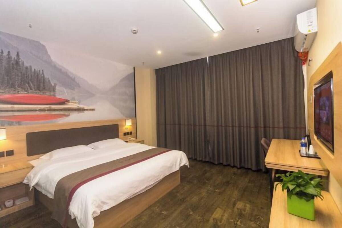 A hotel room with a bed, a desk, a chair, a plant, and a mural of a mountainous landscape.