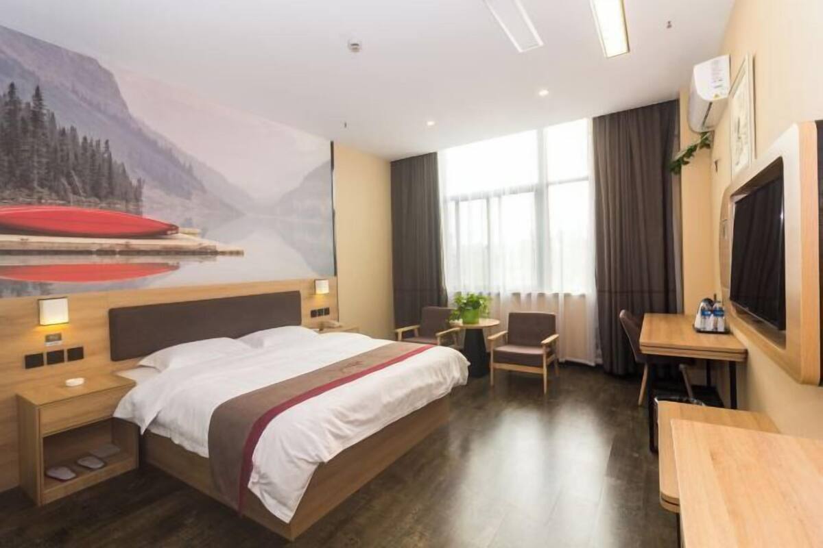 A hotel room with a large bed, a desk, and a chair. There is a window with curtains and a painting of a mountain landscape on the wall.