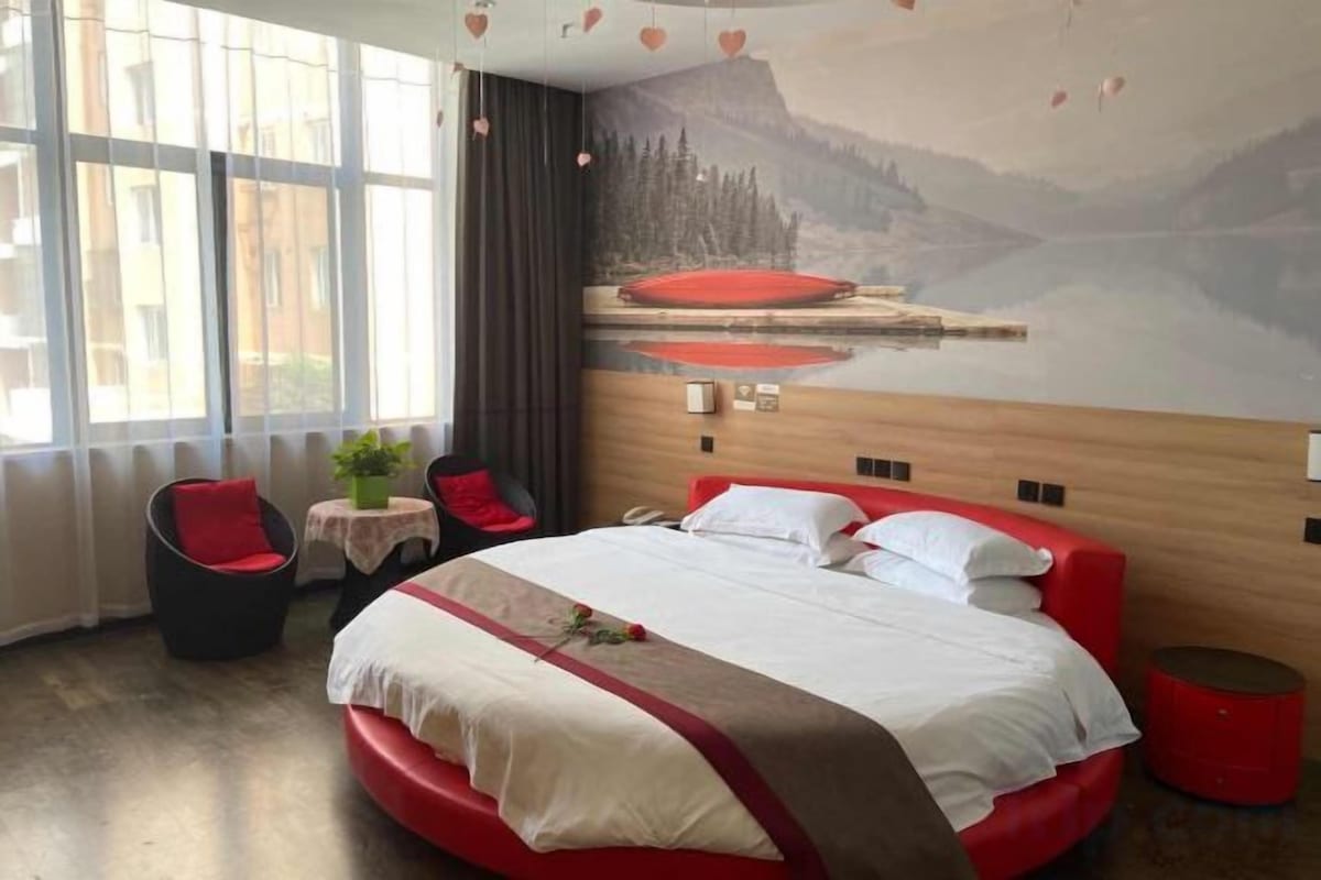 A hotel room with a large bed, a small table with a plant, a red chair, and a view of a mountain landscape through the window.