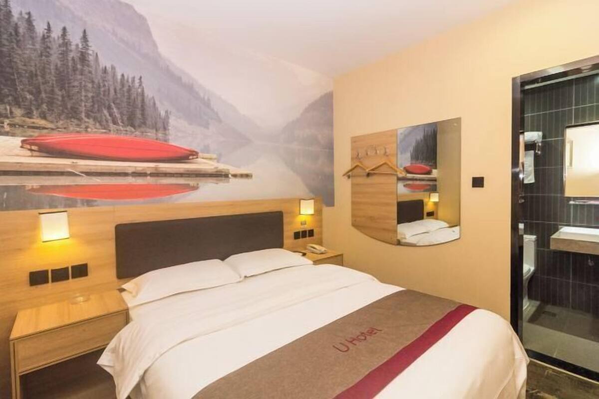 A hotel room with a bed, a nightstand, a mirror, and a wall mural depicting a mountainous landscape.