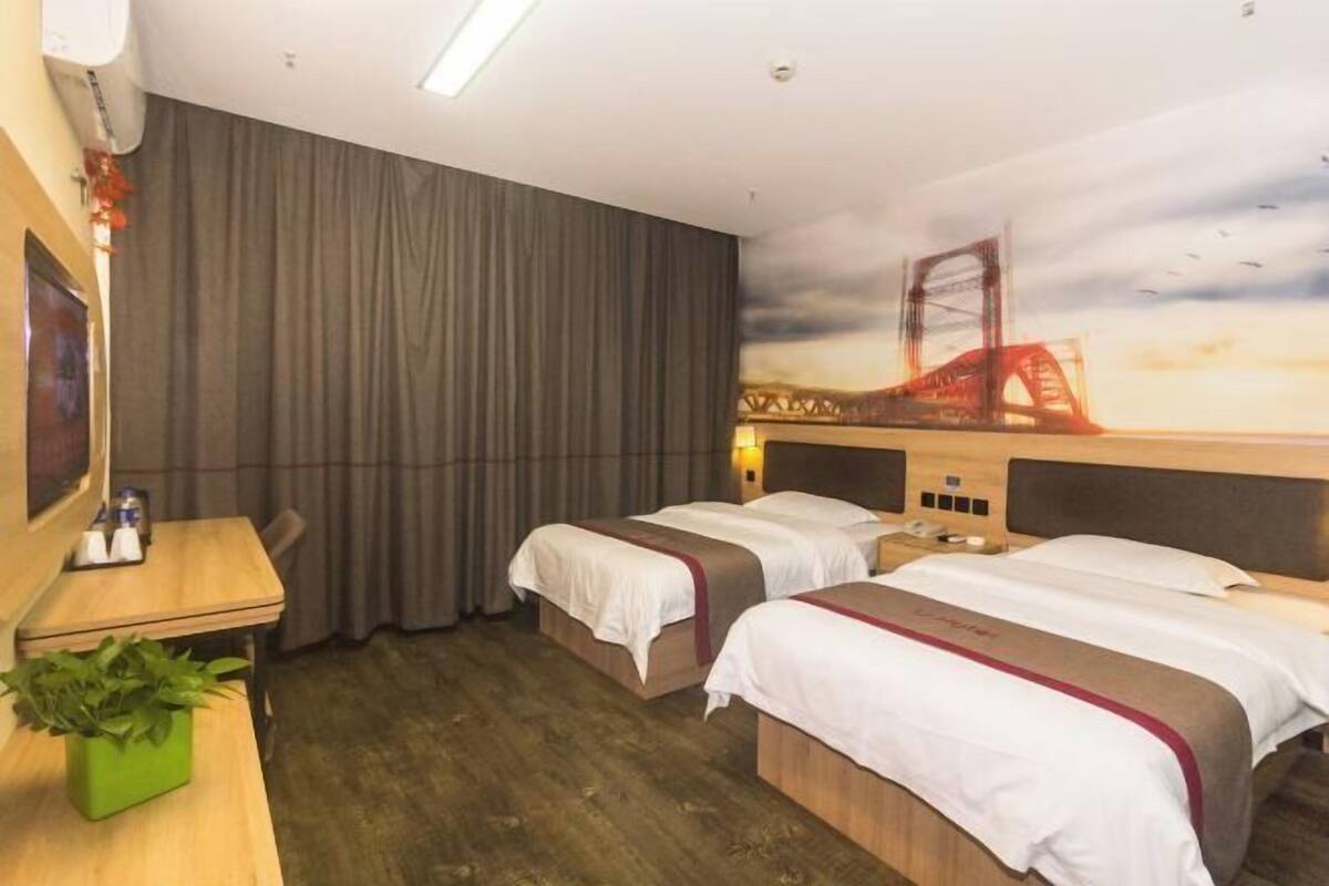 A hotel room with two beds, a desk, a chair, a plant, and a mural of a bridge.