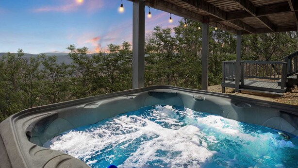 A hot tub with a wooden deck and string lights, surrounded by trees and a view of distant hills. - Mountain cabin w/ hot tub & game room (Romney)