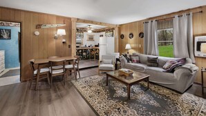 A cozy living room with a wooden floor, a sofa, a coffee table, and a dining area with a table and chairs. - Wood Stove, Fire Pit, Kayaks - 2 Min Walk to River (Springfield)