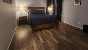 Een slaapkamer met een houten vloer, een bed met een donkerblauwe dekbedovertrek, twee bedlampjes, een plafondventilator en een ingelijst schilderij aan de muur.