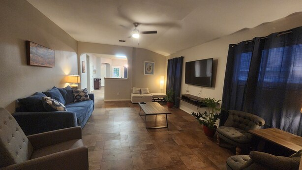 Een woonkamer met een bank, fauteuils, een salontafel en een televisie. Er is een plafondventilator, een lamp en een plant. De vloer is betegeld en er is een deur naar een andere kamer.
