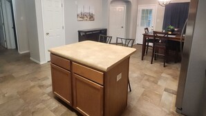 Een keuken met een centraal eiland, houten kasten, een eethoek met stoelen en een tafel, en een koelkast.