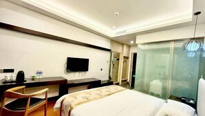 Una habitación de hotel con cama, escritorio, silla, televisor y una pared divisoria de vidrio.