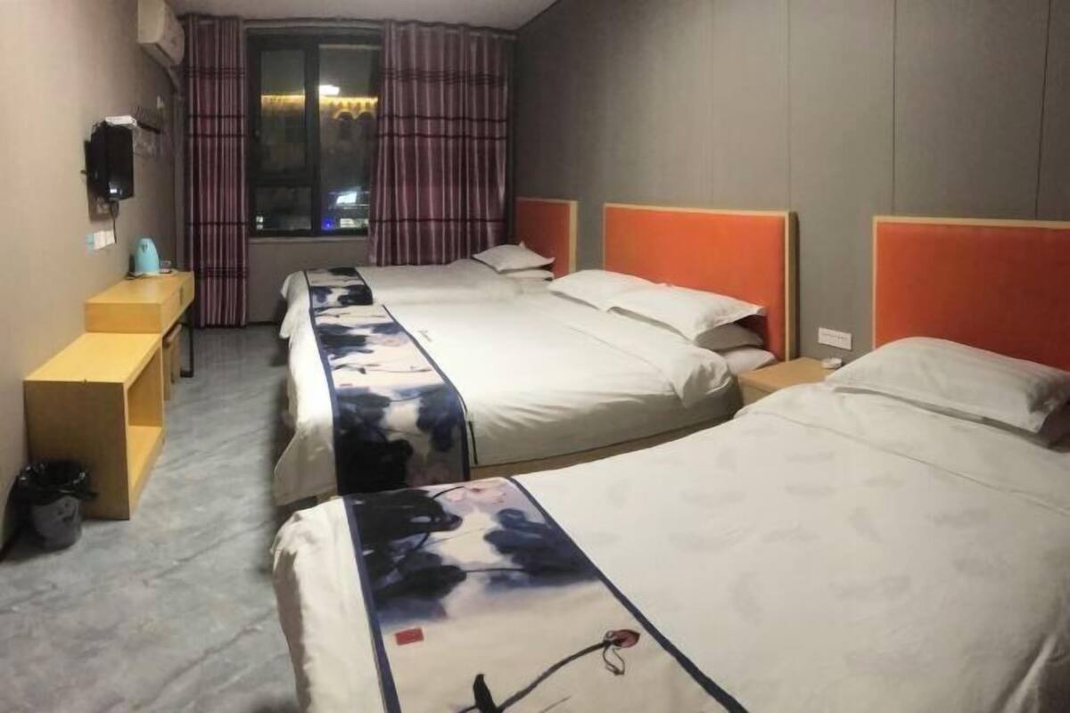 Une chambre d’hôtel avec trois lits, un bureau, une chaise, une fenêtre avec des rideaux et une petite table avec une lampe.