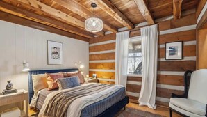 Ein gemütliches Schlafzimmer in einer Blockhütte mit einem großen Bett, einem Sessel, einem Fenster mit Vorhängen und einer Hängelampe.