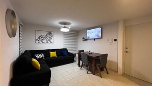 A living room with a black sofa, a dining table with chairs, a ceiling fan, and a TV mounted on the wall. - Nice, cozy, familiar and centrally located apartment (San Pedro Tlaquepaque)