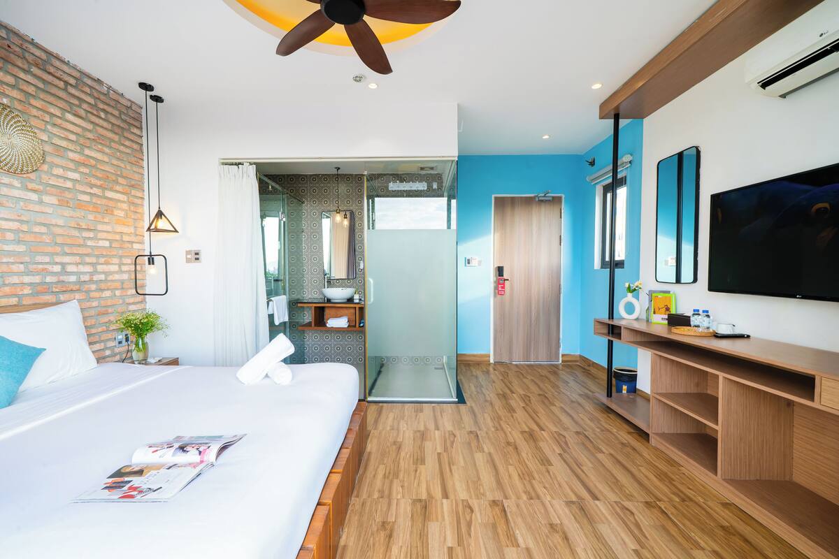 A modern hotel room with a large bed, a TV, a wooden desk, and a bathroom with a glass shower.