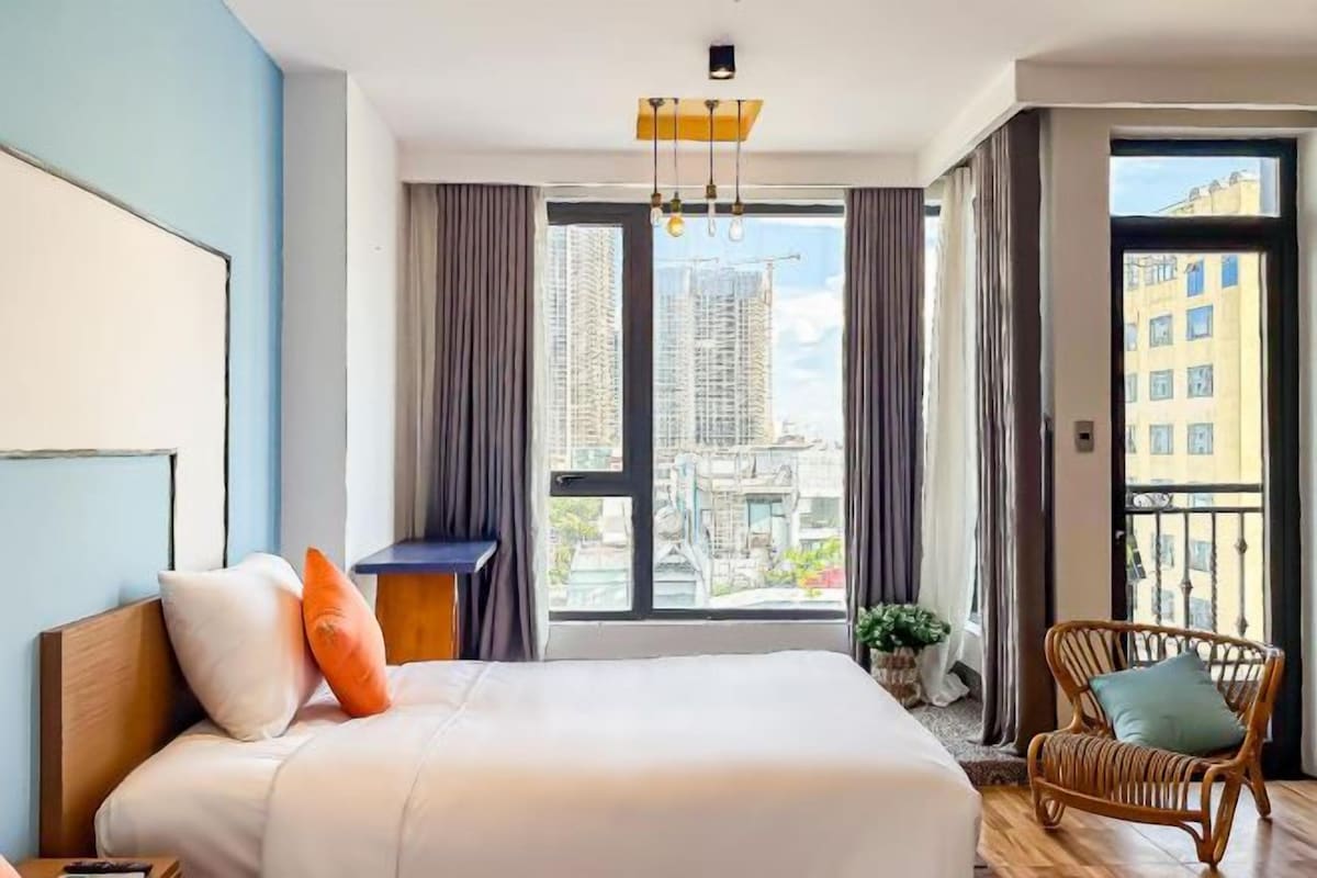 A hotel room with a large bed, a chair, a desk, and a window with a city view.