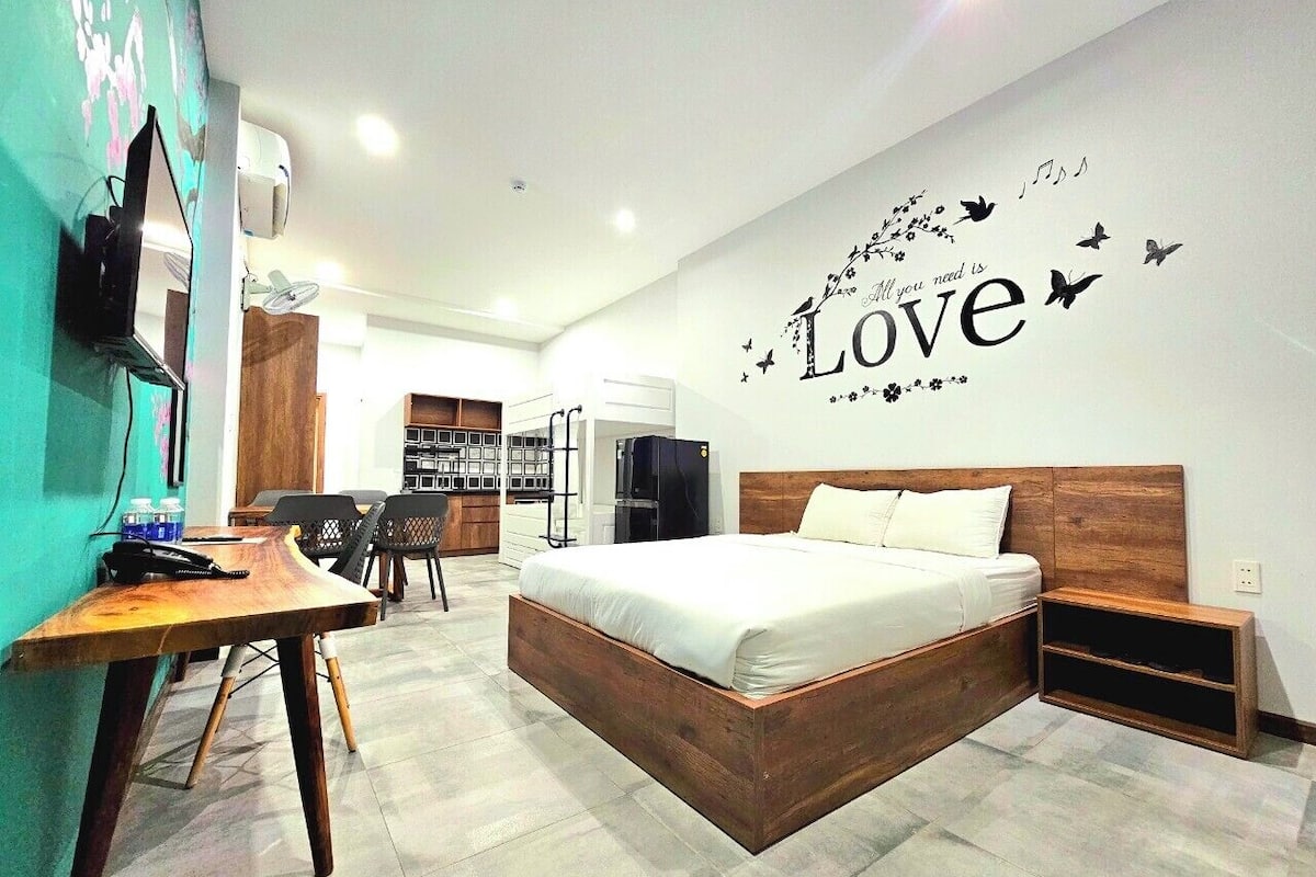 Habitación de hotel moderna con una cama de madera, un escritorio con silla, un armario y una pared decorada con la palabra 'Love'.