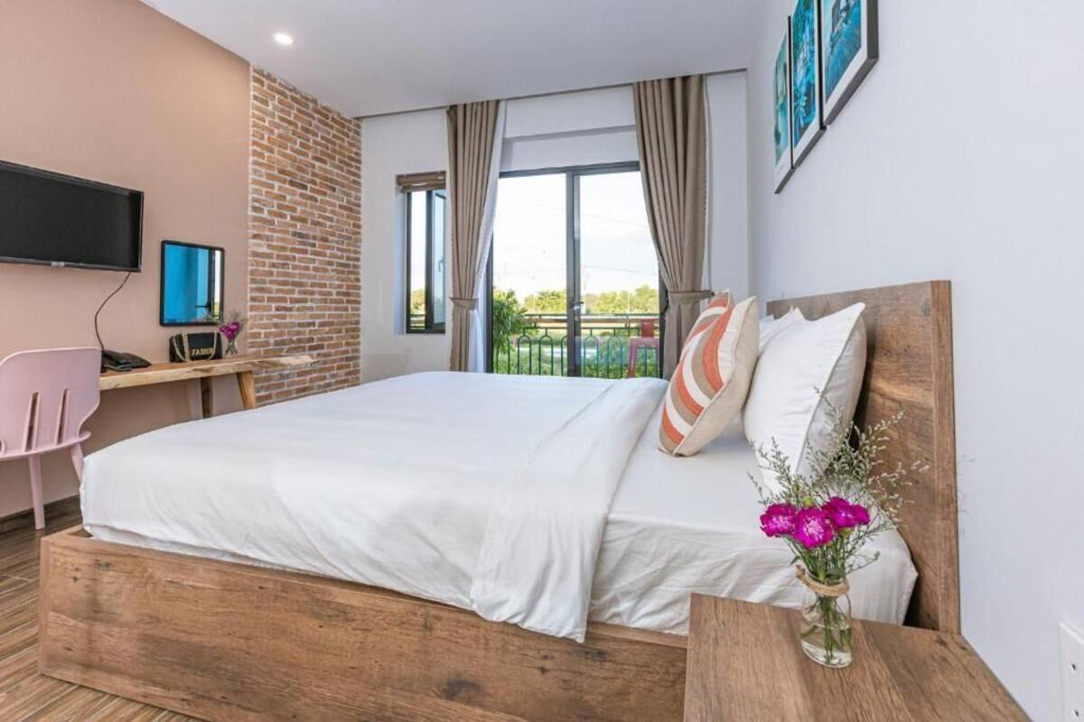Habitación de hotel moderna con una cama de madera, un escritorio con una computadora, un televisor montado en la pared y un ventanal con cortinas.