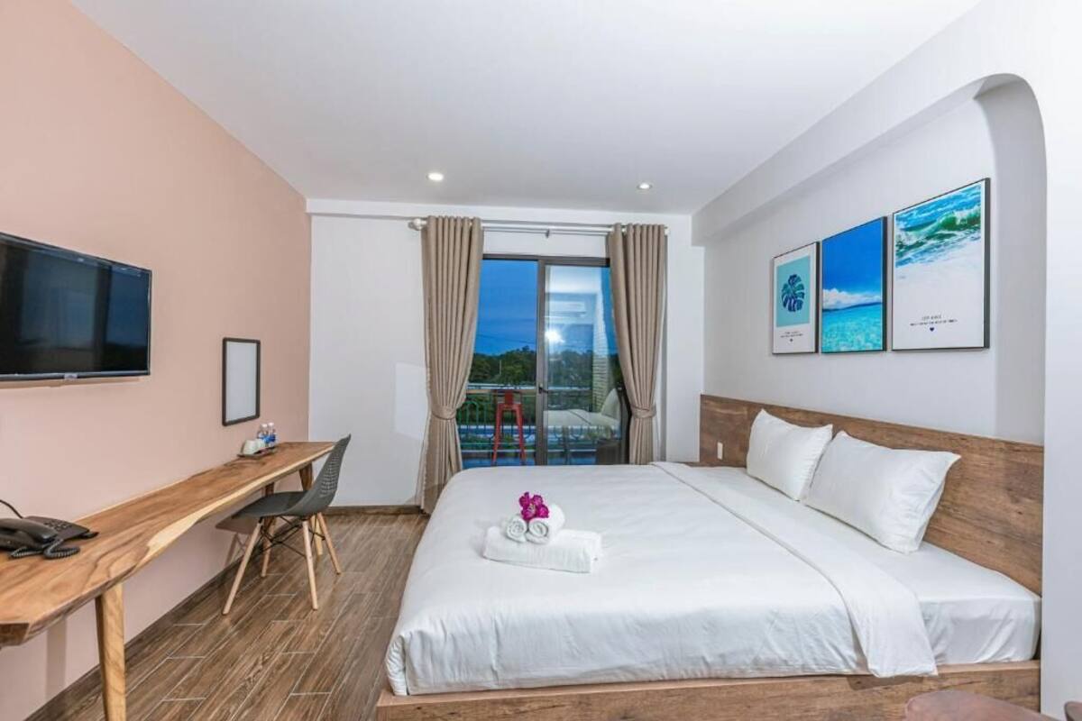 Habitación de hotel con cama, escritorio, silla, televisor y balcón con cortinas.