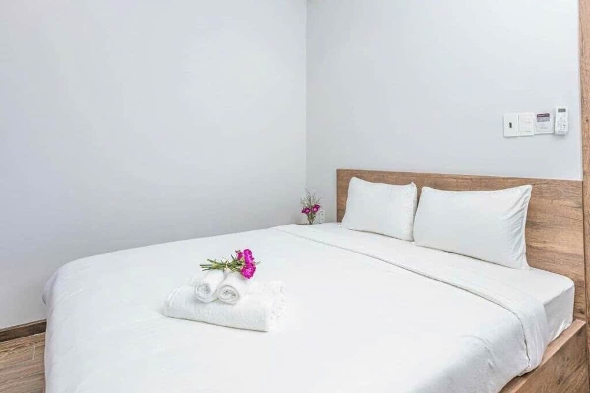 Una cama bien hecha con sábanas blancas, cabecera de madera y una mesita de noche con un florero.