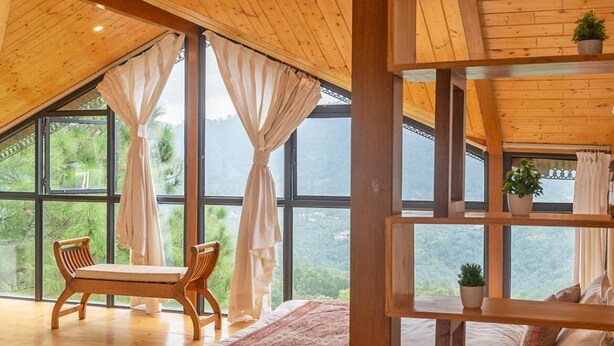 A bedroom with a wooden ceiling, large windows, a bed, a bench, and a shelf with plants. - The Good Vibes by Aero Stays (Kasauli)