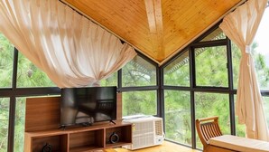 A room with a wooden ceiling, a television on a wooden stand, a window with curtains, and a view of greenery outside. - The Good Vibes by Aero Stays (Kasauli)