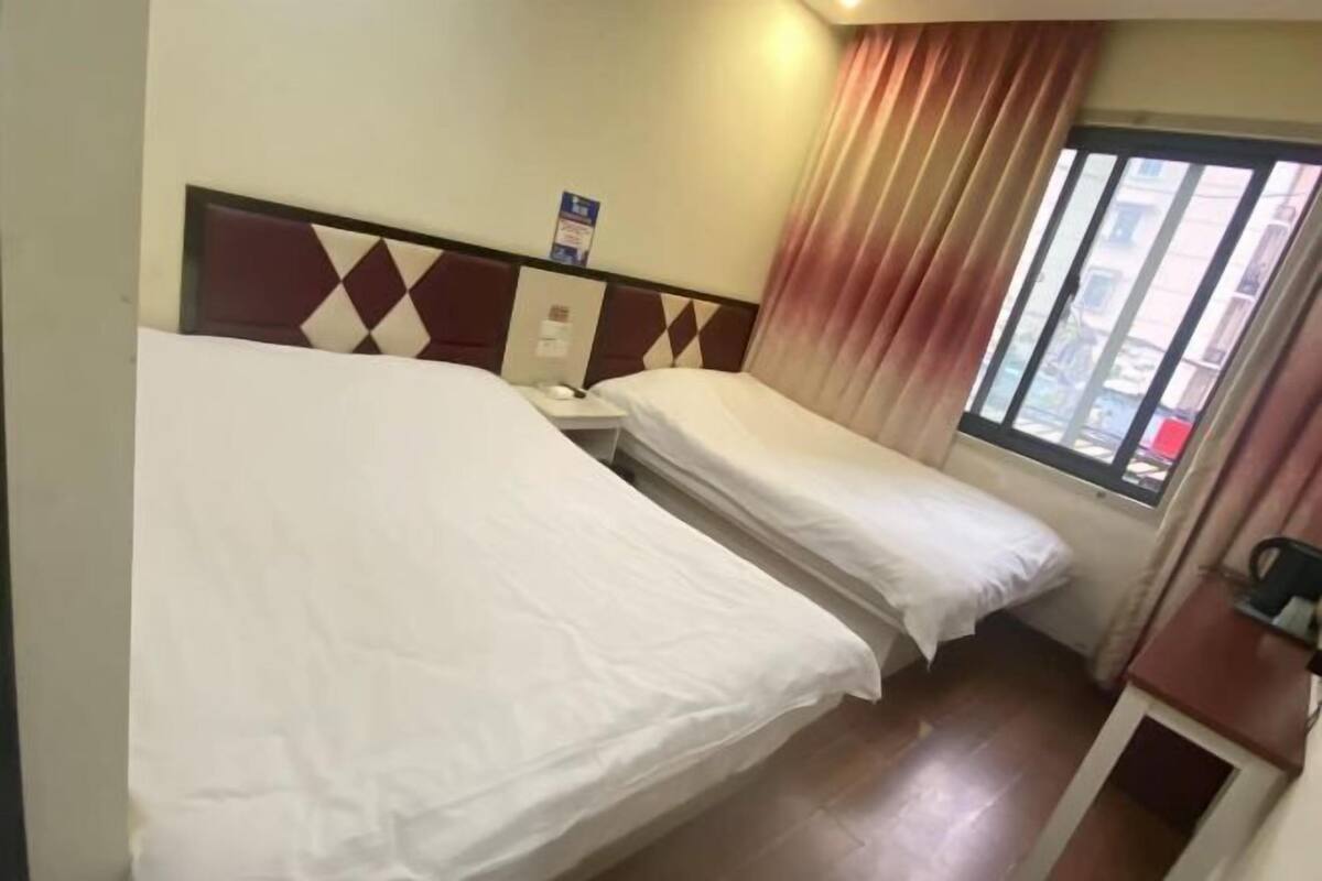 A hotel room with two single beds, a window with curtains, a small desk, and a wall-mounted bulletin board.
