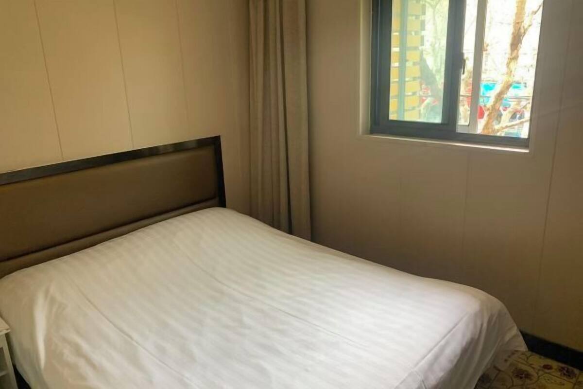 A single bed with a wooden headboard in a room with a window, curtains, and a view of the outside.