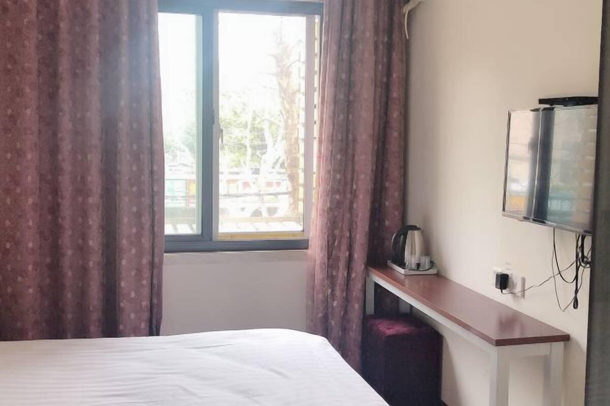 A hotel room with a bed, a desk with a kettle, a TV, and a window with curtains.