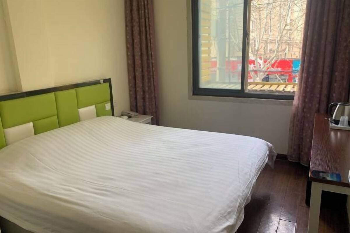 A neatly made bed in a room with a window, a bedside table, and a view of the outside.