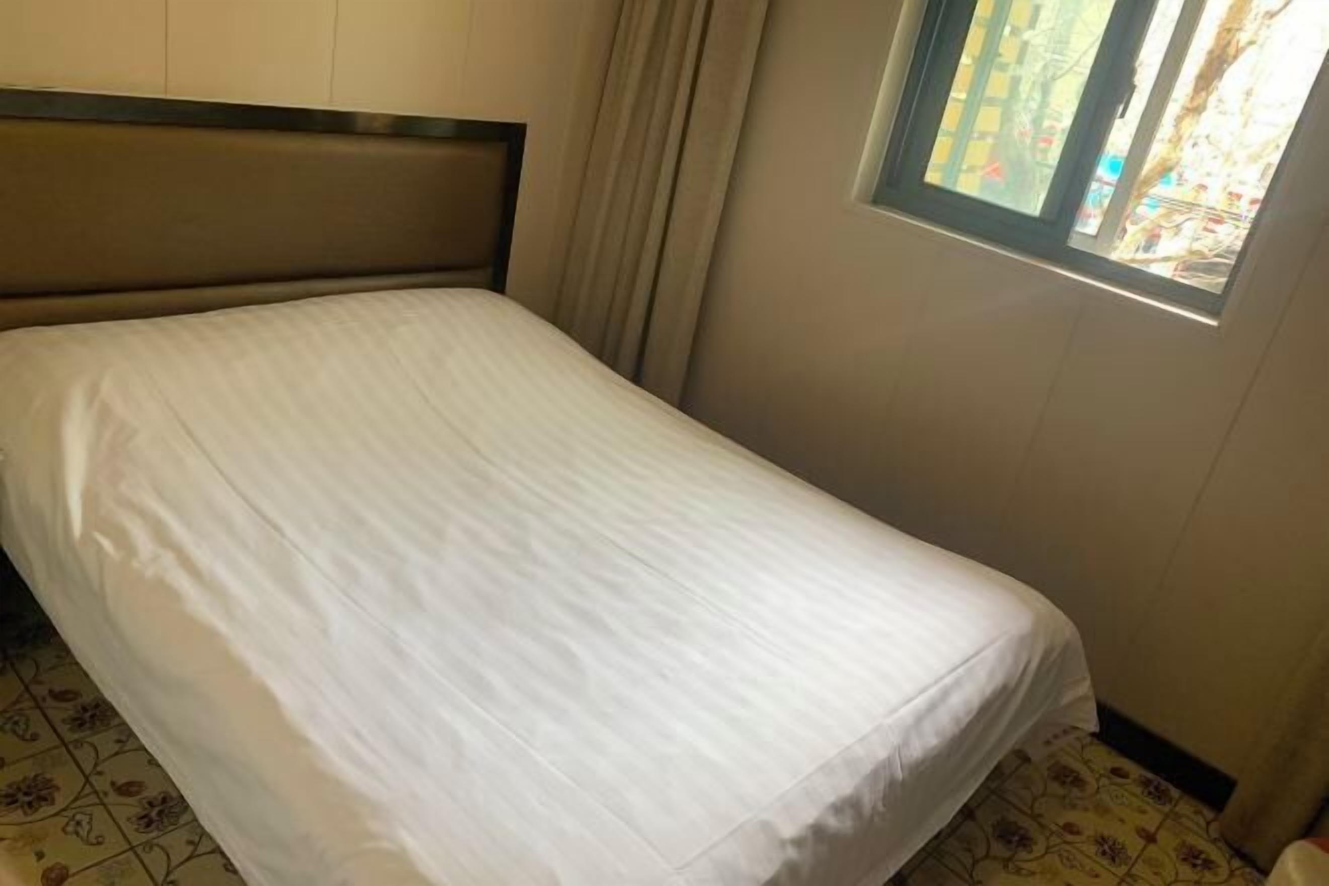 A single bed with a headboard, a window with curtains, and patterned flooring.