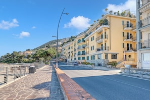 A coastal street with multi-story buildings, a clear sky, and a stone-paved sidewalk.