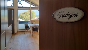 Un interior moderno con un letrero de madera que dice "Hochgern" en una puerta, una zona de comedor con dos sillas y una mesa, y vistas a través de ventanas amplias.
