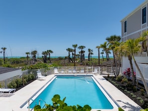 A rectangular swimming pool with a clear blue water, surrounded by a white fence. There are several lounge chairs and a small deck area. Palm trees and other vegetation are visible in the background, with a view of the ocean and a clear sky.