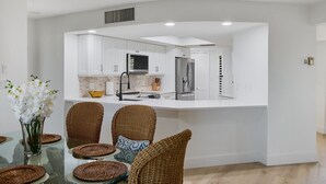 Une cuisine moderne avec des meubles blancs, un îlot central et un coin repas agrémenté de chaises en osier et de sets de table tressés.