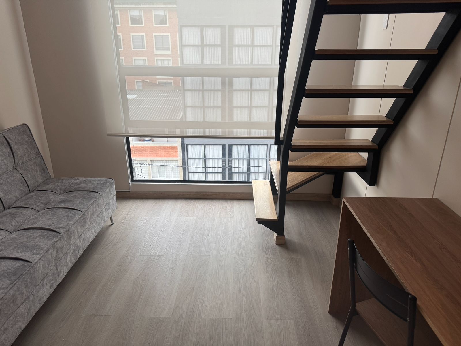 A room with a grey sofa, a wooden desk, and a black staircase.