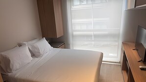 A neatly made bed with white linens, a wooden nightstand, a television, and a window with blinds. - Vivo52 (Bogotá)