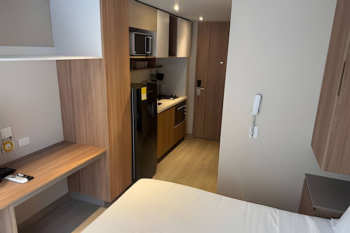 A compact hotel room with a bed, a desk, a kitchenette, and a phone on the wall.