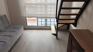 A modern loft bedroom with a grey sofa, a wooden desk, and a black staircase. - Vivo52 (Bogotá)