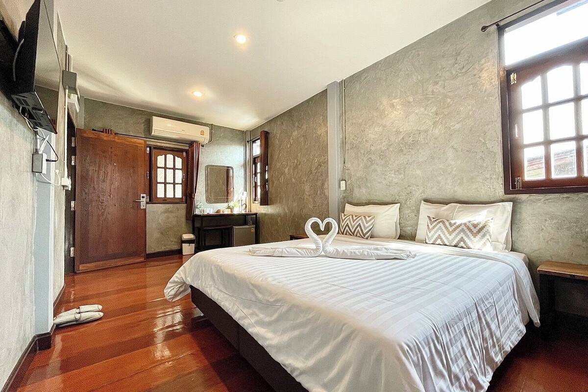 A bedroom with a large bed, wooden flooring, a wooden wardrobe, a mirror, and a window with curtains.