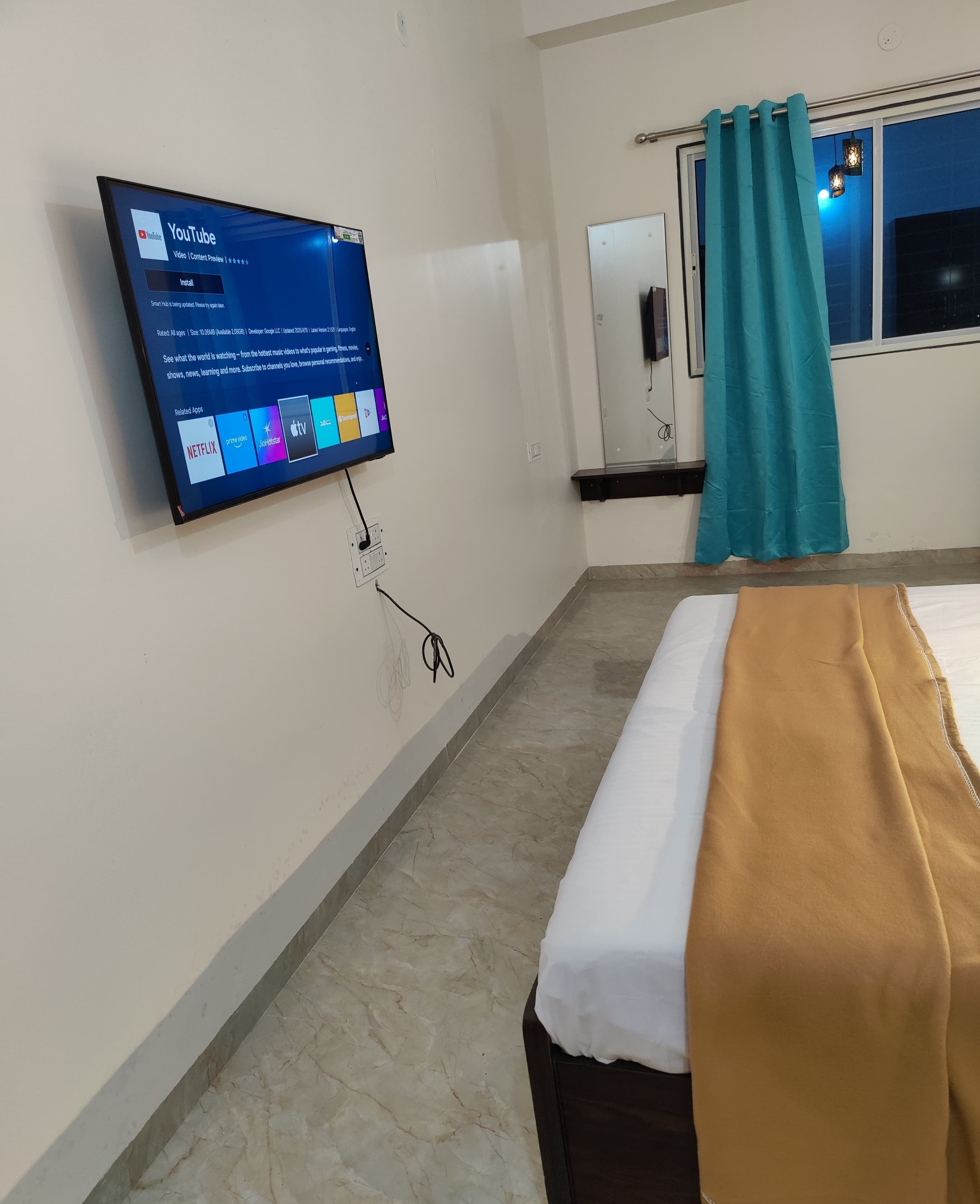 Habitación de hotel con cama, televisor montado en la pared mostrando YouTube, ventana con cortina y una pequeña mesa con lámpara.