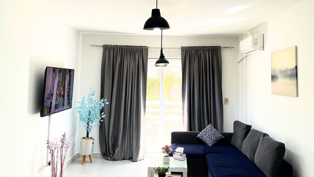 Een moderne woonkamer met een donkerblauwe bank, een klein bijzettafeltje en een groot raam met grijze gordijnen. Daarboven hangt een zwarte hanglamp boven de bank en aan de muur hangt een lijst met een afbeelding.